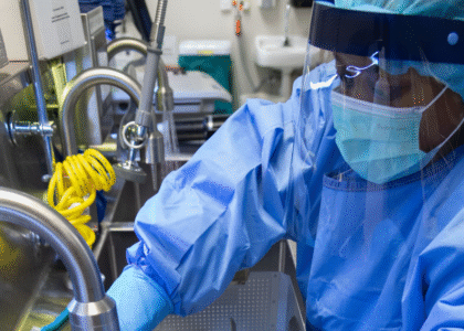 A sterile processing technician wearing full PPE, including a face shield and surgical mask, performs instrument cleaning at a decontamination sink. This image represents critical steps in safe reprocessing and workplace safety aligned with AAMI ST58:2024 guidelines.