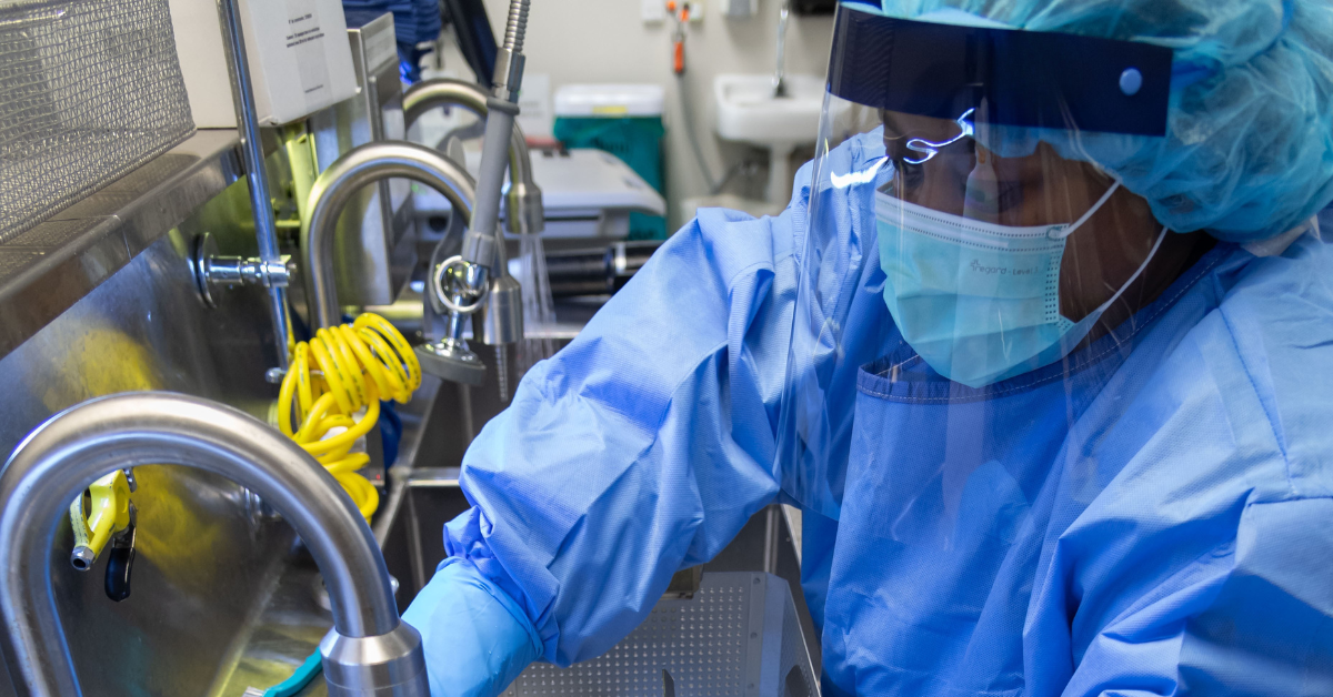 A sterile processing technician wearing full PPE, including a face shield and surgical mask, performs instrument cleaning at a decontamination sink. This image represents critical steps in safe reprocessing and workplace safety aligned with AAMI ST58:2024 guidelines.