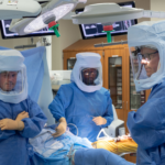 Three operating room clinicians wearing powered air-purifying respirators (PAPRs) during a surgical procedure, demonstrating the specialized professionals trusted by hospital staffing companies.