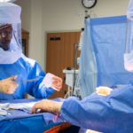 Operating room clinicians wearing powered air-purifying respirators (PAPRs) during a surgical procedure in a hospital operating room.